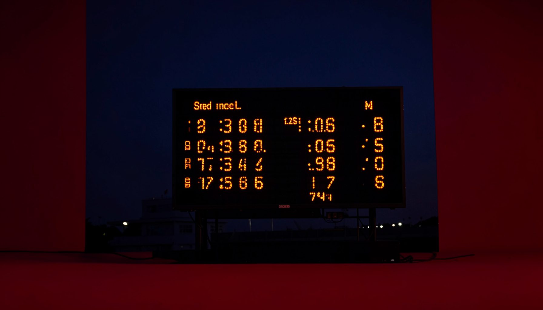Stadium scoreboard at dusk showing cricket match figures, deep navy sky with warm orange digits